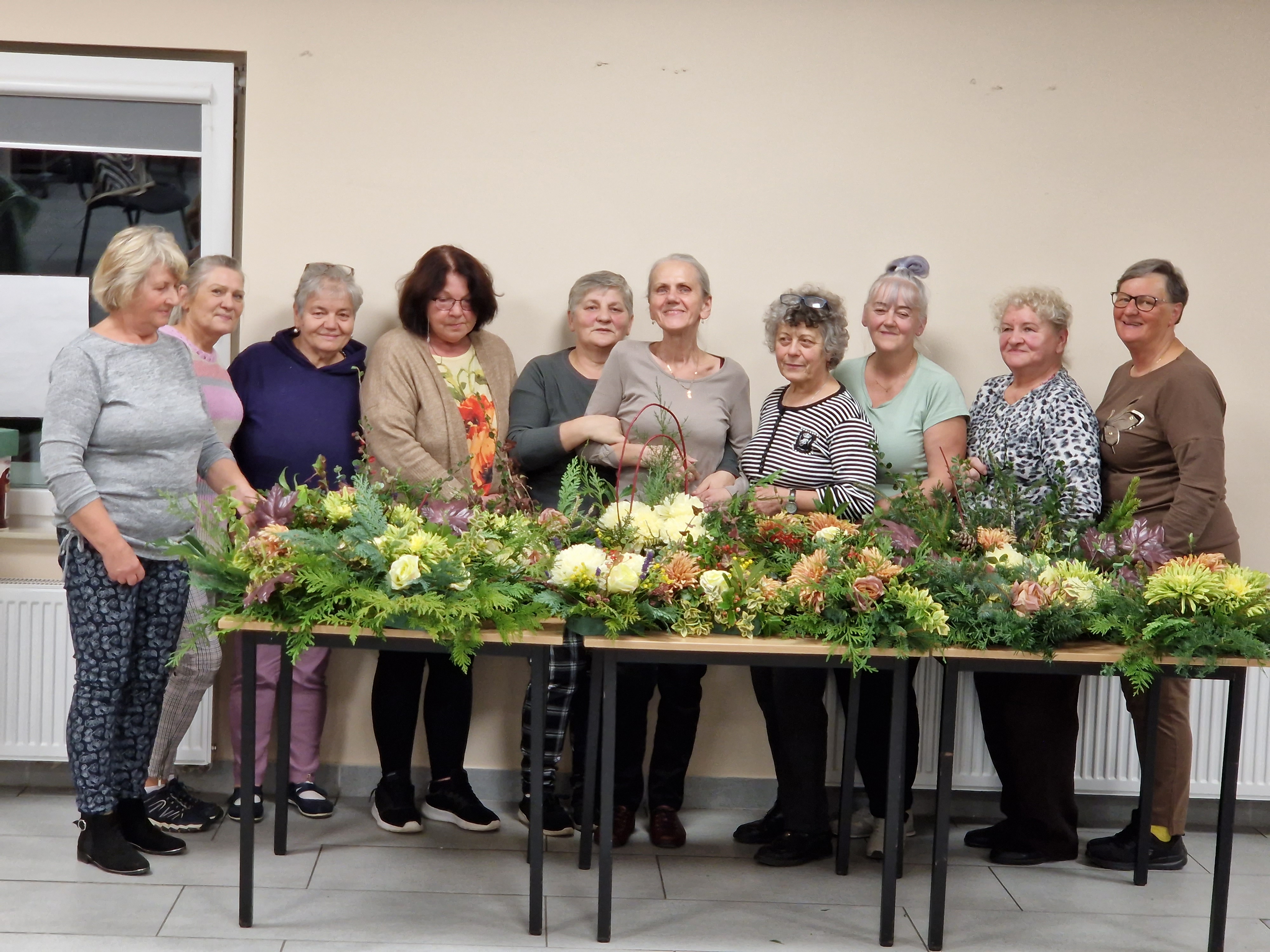 Warsztaty florystyczne w Klubie Seniora w Widzinie - uczestnicy warsztatów stoją na tle wykonanych stroików florystycznych